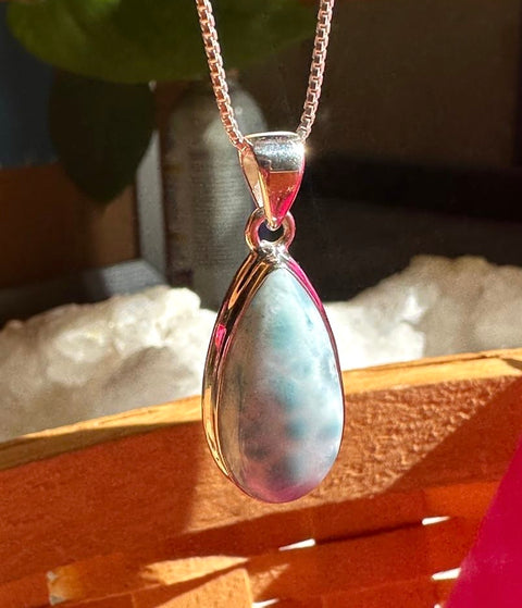 Teardrop-shaped pendant with a pink and white marbled design on a chain, set against a blurred natural background.